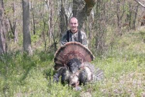 10-1-scaled A man poses with his turkey in the woods.