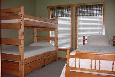 Bedroom with a wooden bunk bed and a separate wooden twin bed, white bedding, and two windows with plaid valances and white blinds.