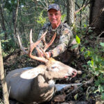 Man in camouflage clothing and cap kneeling in a wooded area behind a harvested white-tailed buck with large multi-point antlers, holding the antlers with one hand.