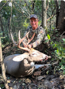 BF_Man_with_deer_2025 Man in camouflage clothing and cap kneeling in a wooded area behind a harvested white-tailed buck with large multi-point antlers, holding the antlers with one hand.