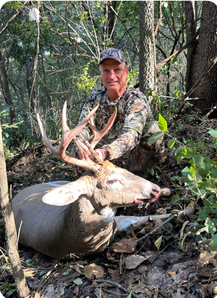 Man in camouflage clothing and cap kneeling in a wooded area behind a harvested white-tailed buck with large multi-point antlers, holding the antlers with one hand.